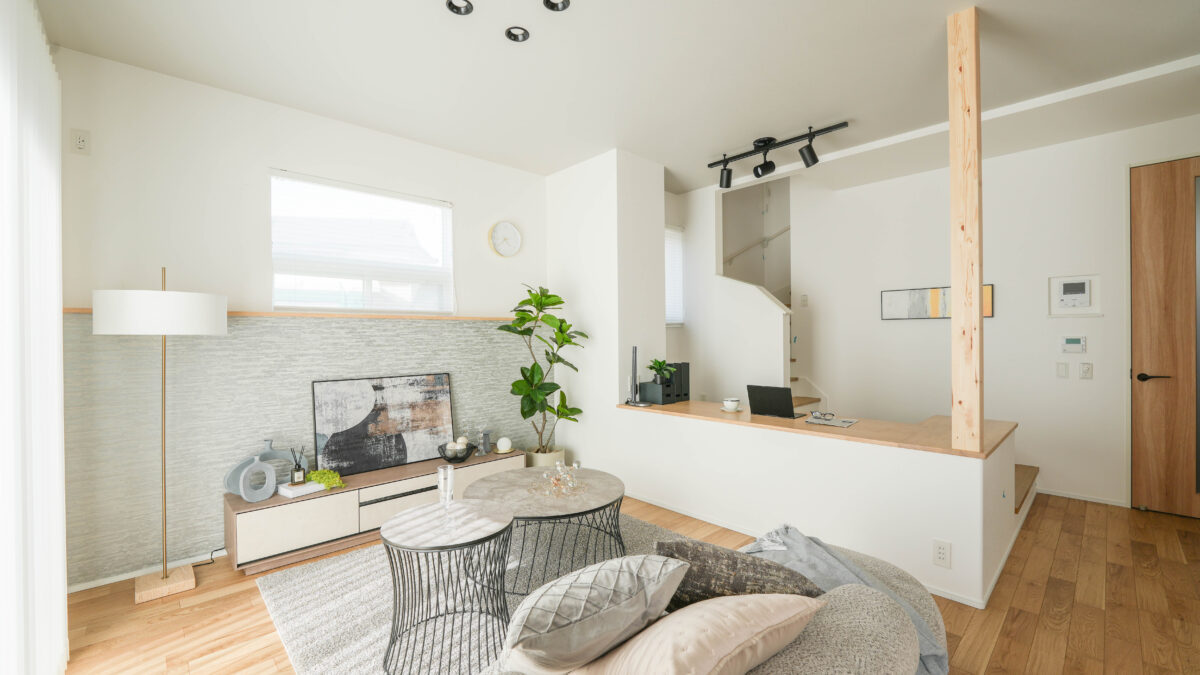 白い壁と木目フローリングのリビング空間。左に窓とフロアランプ、中央にソファと2つの丸いテーブル、奥にテレビボードと観葉植物、右側にカウンターと階段、天井にはスポットライトが設置されている。