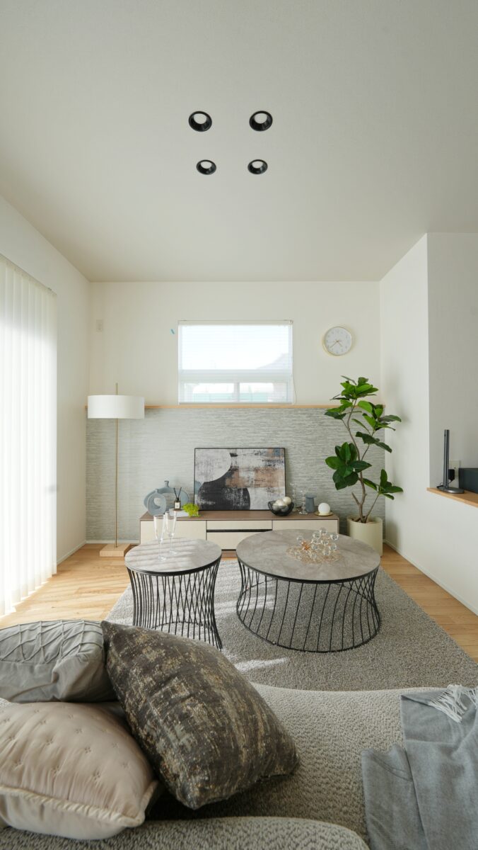 明るい自然光が差し込む、シンプルで洗練されたリビングルームの様子。中央には丸いローテーブルが2つ並び、その上にはガラスの装飾品やグラスが置かれている。奥にはローボードがあり、抽象画や花瓶、小物がバランスよくディスプレイされている。右側には観葉植物が配置され、空間にやさしいアクセントを加えている。白を基調とした壁と天井に、木目のフローリングやモダンな家具が調和し、落ち着いた居心地のよい空間を演出している。