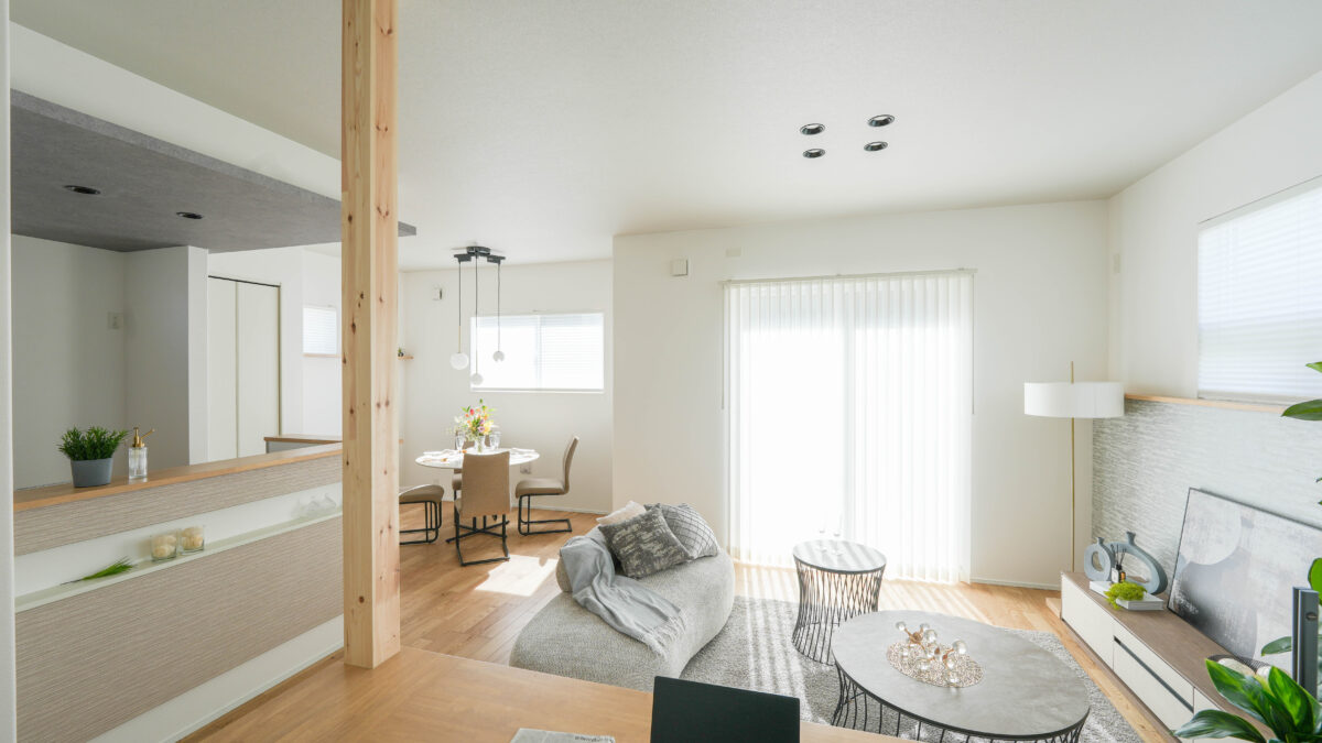 白い壁と木目フローリングのリビングダイニング空間。中央にソファと2つの丸いテーブル、右側にテレビボードとフロアランプ、左奥にダイニングテーブルと椅子が配置されている。大きな掃き出し窓から自然光が入り、左手にはキッチンカウンターと木の柱が見える。