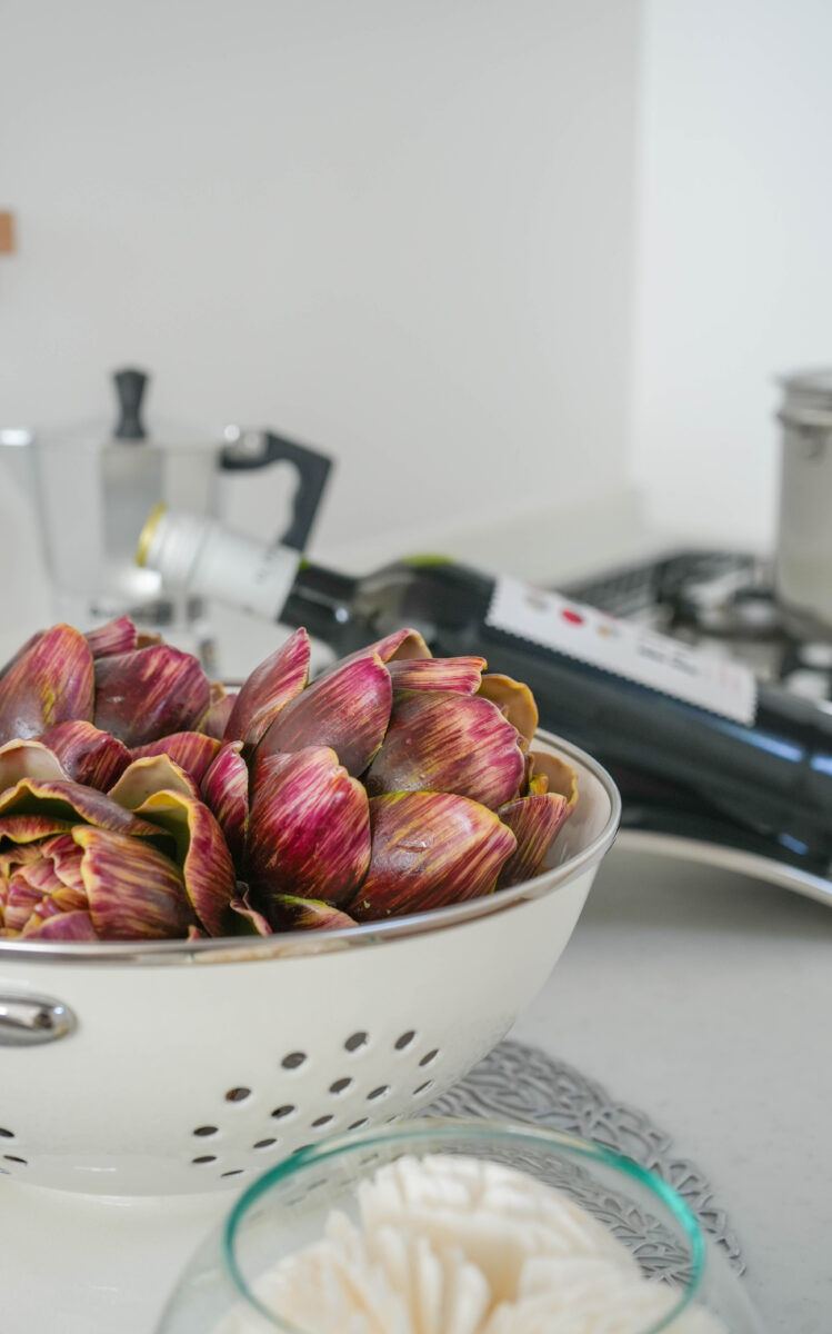 キッチンカウンターに置かれた白いボウルに、紫がかった皮のアーティチョークがたっぷりと盛られている様子。奥にはコーヒーメーカーや調理器具が並び、日常の調理風景を感じさせる。落ち着いた色合いの中でアーティチョークの鮮やかな色味が引き立っている。