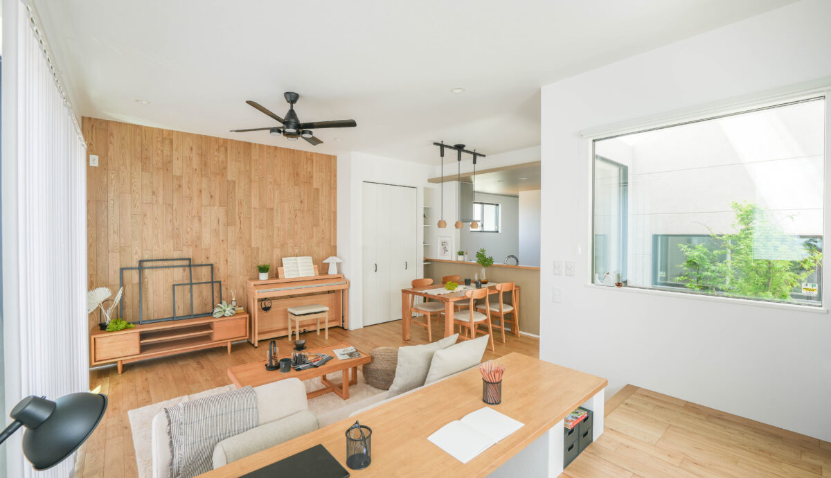 フロアを貼り上げた壁がアクセントとなっているLDK。高い天井にシーリングファンが、ダイニングにはマットな質感のベージュ色のペンダントライトが3灯ついて、どちらもブラックなため統一感のあるコーディネート。オーク色のピアノとテレビボードがぴったりのサイジングで配置されている。