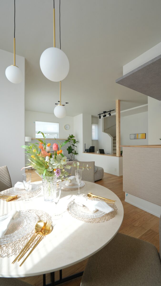 自然光がたっぷりと差し込むダイニングキッチンの様子。丸いダイニングテーブルにはガラスの花瓶に生けられた色とりどりの花が飾られ、繊細なプレートやグラス、ゴールドのカトラリーが上品にセッティングされている。上部には球体のペンダントライトが吊り下げられ、空間に柔らかなアクセントを添えている。奥にはリビングスペースや階段が見え、開放感のある間取りが広がる。白を基調とした内装に木目の床や家具が調和し、明るく洗練された暮らしのシーンを演出している。