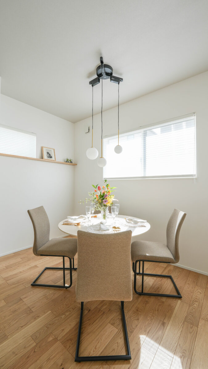 白い壁と木目フローリングのダイニング空間。中央に丸いダイニングテーブルと4脚の椅子が配置され、テーブルには花と食器がセッティングされている。天井からは球体のペンダントライトが3灯下がり、右側の窓から自然光が差し込む。左壁には飾り棚と小さなアートが設けられている。